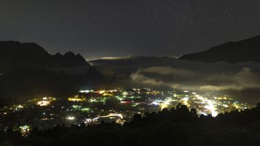 Vue de Cilaos la nuit depuis les au dessus de la ville, les lumières des rues en contrebas éclairent quelques nuages bas dans le noir.