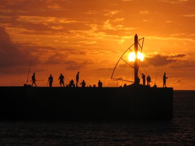 Coucher de soleil orange avec jetée et environ 10 silhouettes noires de pêcheurs qui se découpent sur la mer.