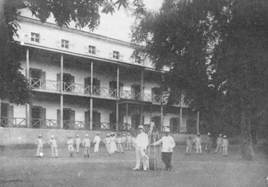 photo noir et blanc d'un parc avec plusieurs personnes vêtues de blanc posent avec de casques coloniaux devant un bâtiment colonial avec des coursives