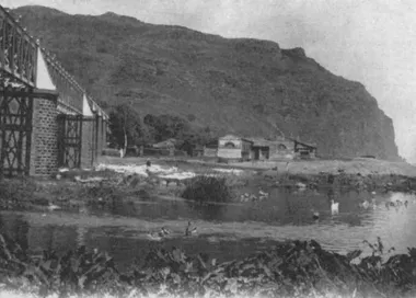 photo noir et blanc d'un bâtiment au pied d'une falaise de l'autre coté d'une rivière