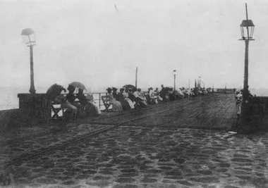 photo noir et blanc d'une jetée avec des gens assis le long avec des ombrelles