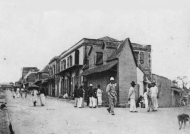 photo noir et blanc d'un angle de rue avec des bâtiments de formes diverses. des personnes déambulent, femme avec ombrelles, hommes avec chapeaux et militaires avec casque colonial