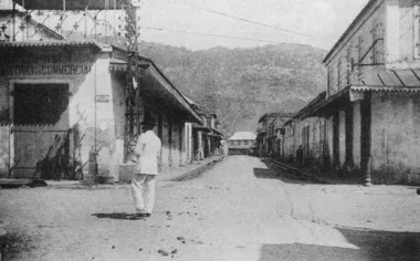 photo noir et blanc d'une rue bordée de maisons rectilignes blanches avec auvent. Une montagne découpe l'horizon et un hommeen blanc se tient sur le devant