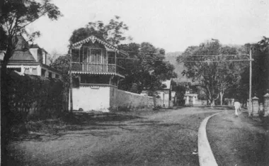 photo noir et blanc d'une rue tournante bordée de murs et d'un large trottoir. Une gueritte a lambrequinssurplombe un mur