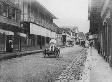 photo noir et blanc d'une rue étroite bordée de maisons avec auvents. Dans la rue roulent un carosse et une voiture automobile