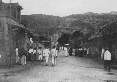 photo noir et blanc d'une rue bordée de maisons basses menant vers une église. De nombreuses personnes vêtues de blanc s'y dirrigent